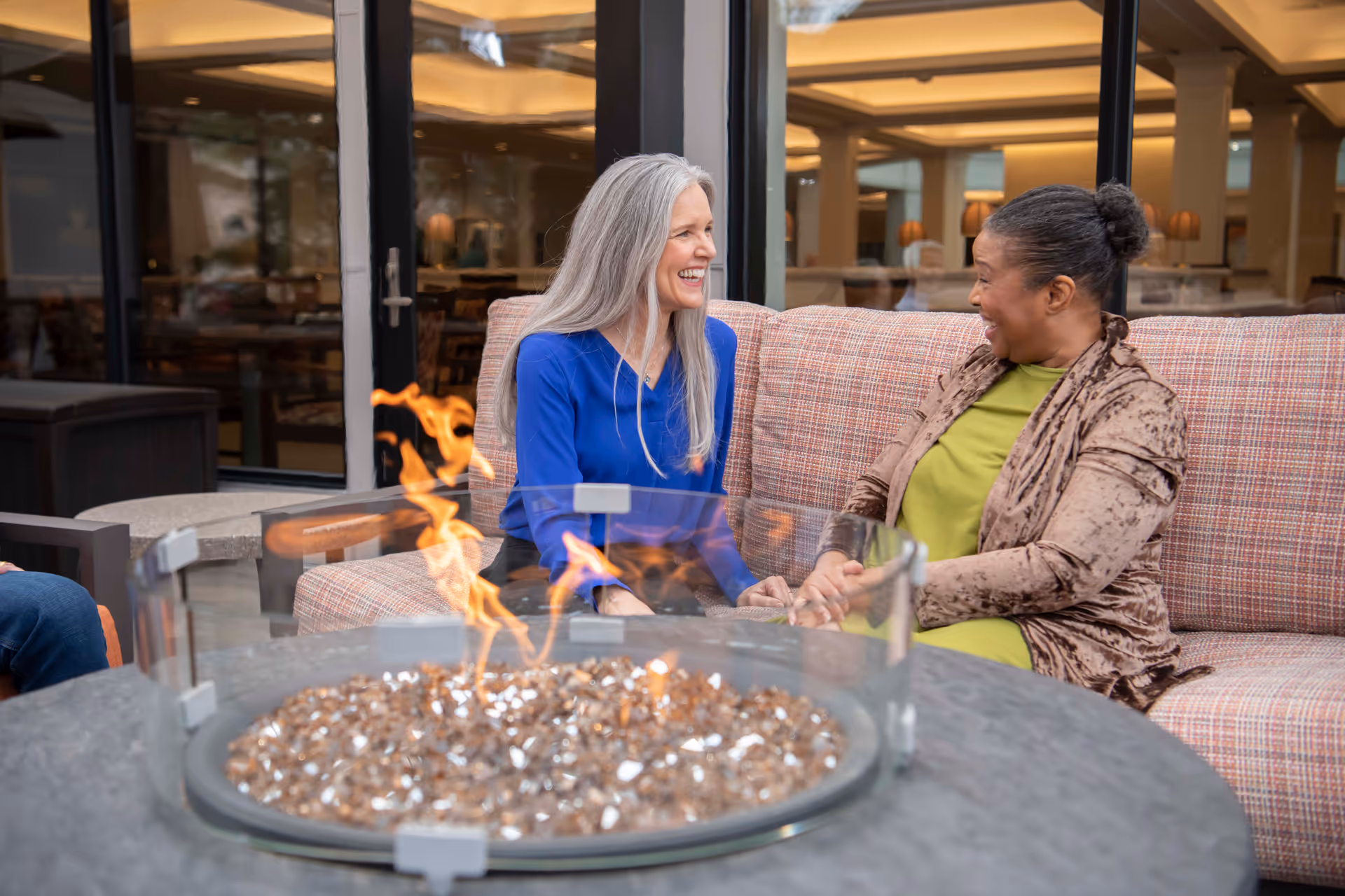 Two women sitting on a cushioned outdoor sofa, smiling and holding hands near a round fire pit with flames and glass wind guard, with a building interior visible through glass doors behind them.