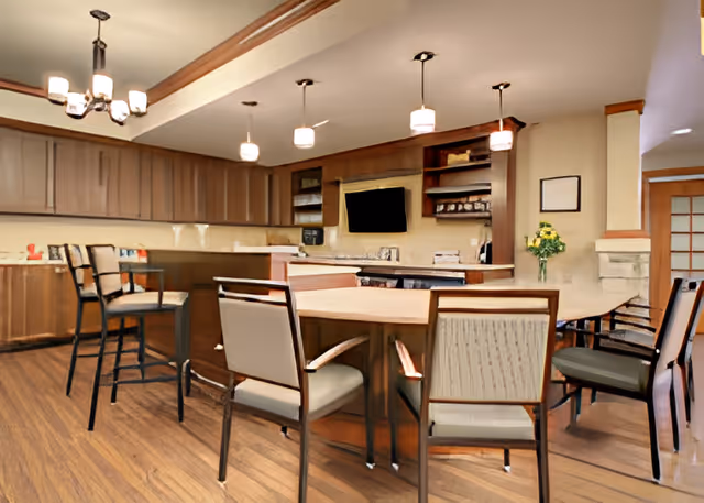 Interior view of a senior living facility's kitchen and dining area with wooden cabinets, a countertop with bar stools, a dining table with chairs, pendant lights, and a wall-mounted TV.