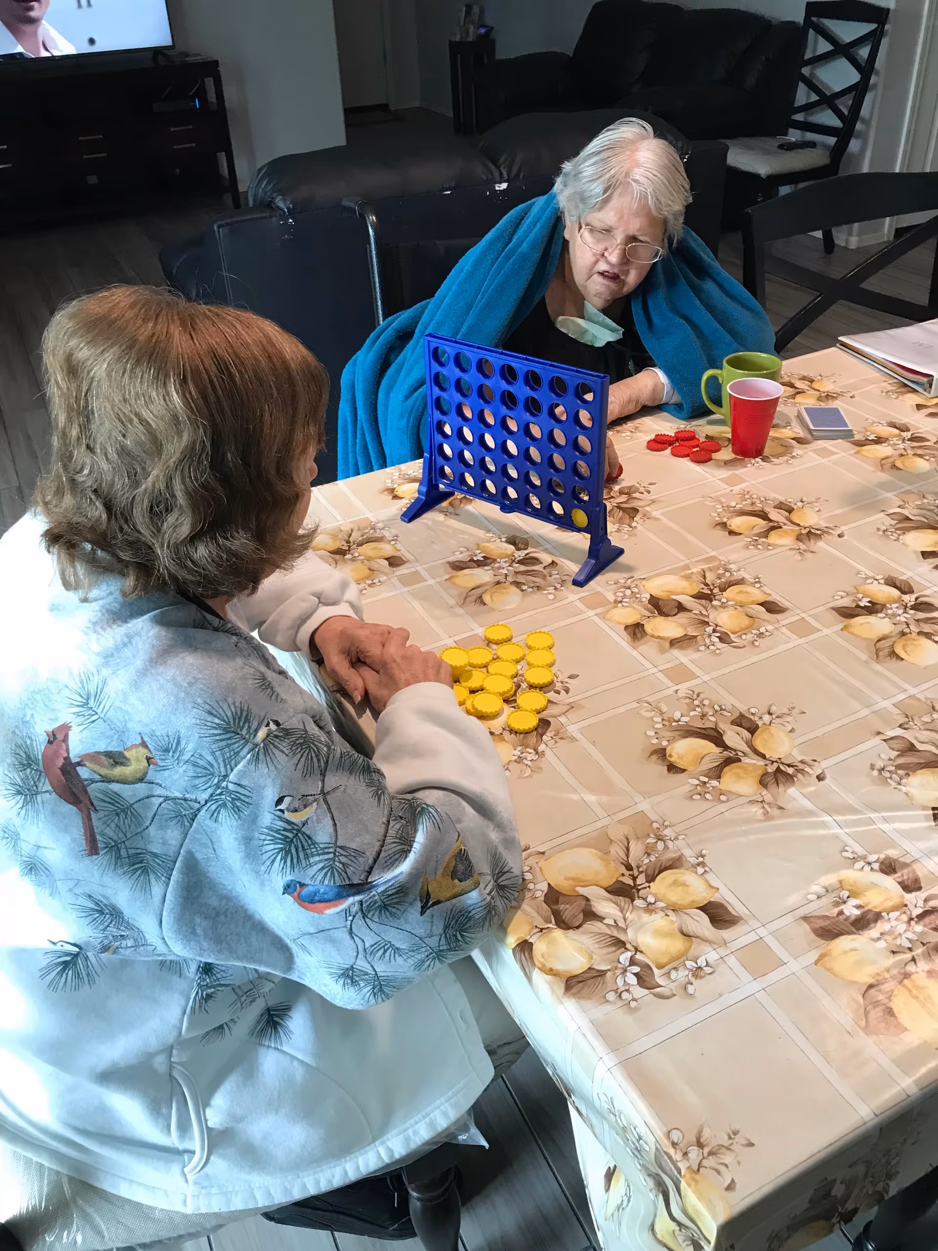 Two elderly women sitting at a table playing a game of Connect Four. One woman is wearing a white jacket with bird designs and holding yellow game pieces, while the other woman is wrapped in a blue blanket and has red game pieces. The table has a floral-patterned tablecloth, and there are cups and papers on the table. A living room with a TV and furniture is visible in the background.
