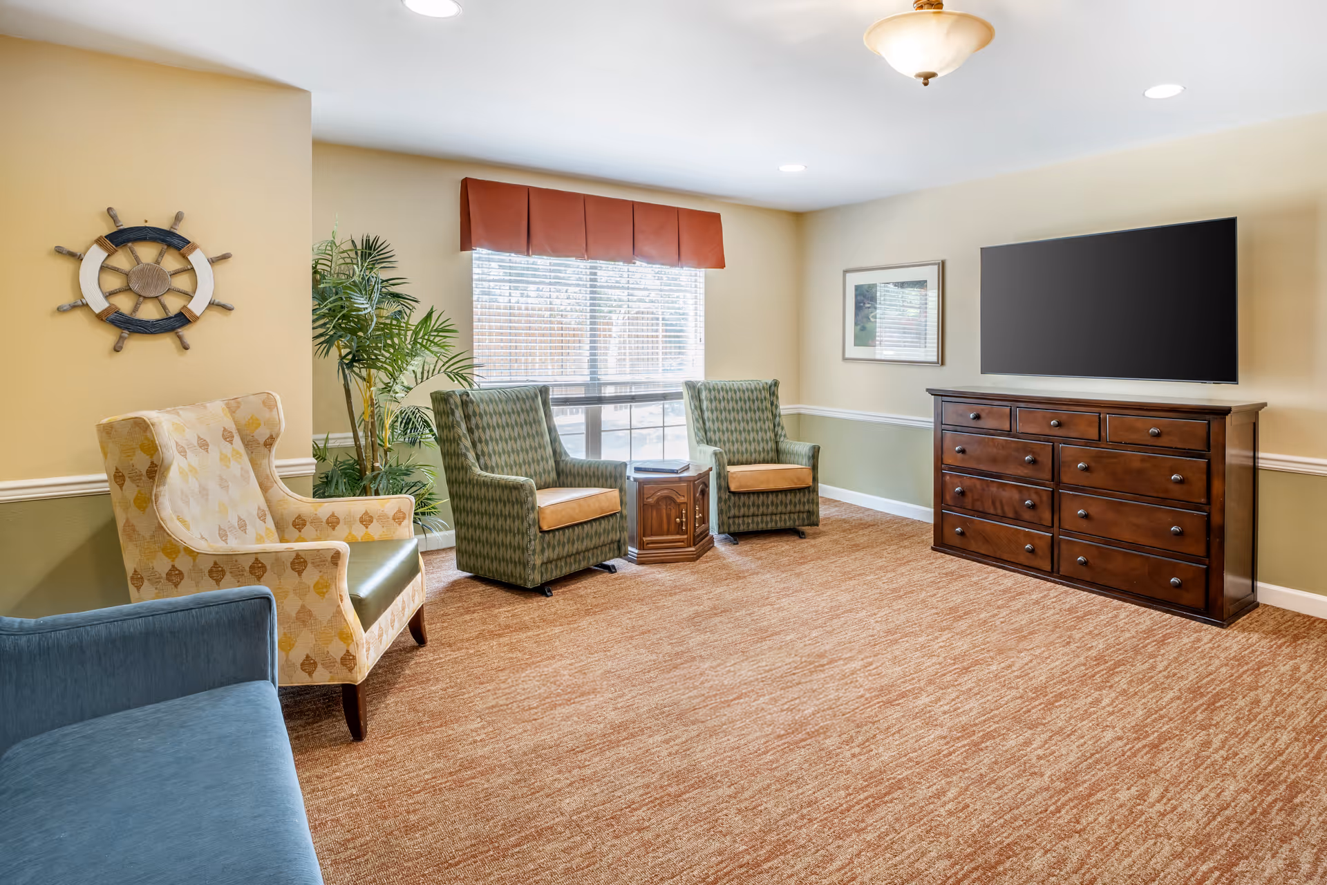 A cozy living room with beige and green walls, a large window with blinds and a red valance, two green patterned armchairs with a small wooden side table between them, a beige patterned wingback chair with a green seat cushion, a blue sofa, a large wooden dresser with multiple drawers, a flat-screen TV mounted above the dresser, a framed picture on the wall, a decorative ship wheel on the wall, and a potted plant near the window.