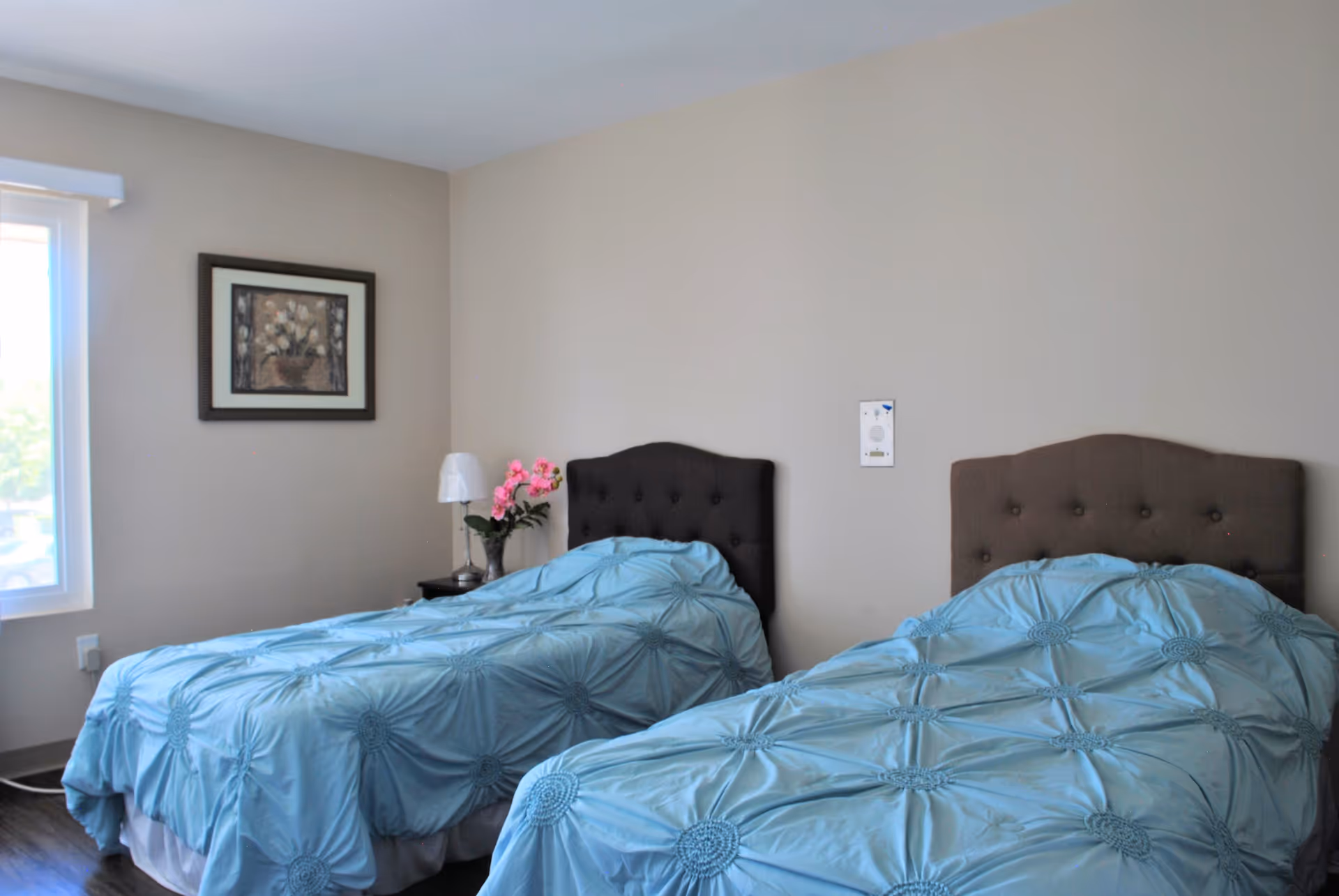 A bedroom with two twin beds covered in light blue bedspreads with a circular pattern. Each bed has a dark upholstered headboard. Between the beds is a small nightstand with a lamp and a vase of pink flowers. A framed floral painting hangs on the beige wall above the nightstand. A window on the left side lets in natural light.