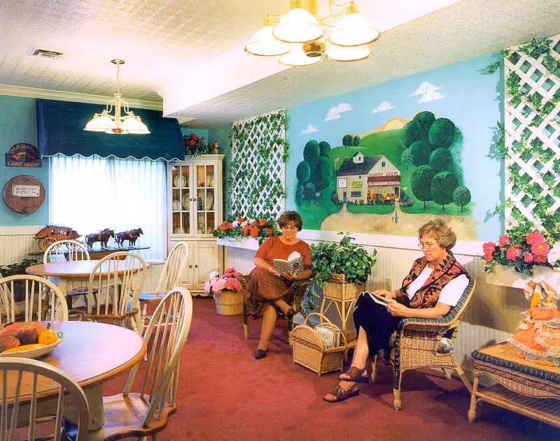 A cozy common area with two elderly women sitting in wicker chairs reading books. The room has round wooden tables and chairs, a cabinet with dishes, and decorative plants. The walls are adorned with a large mural depicting a countryside scene with trees, a house, and a sunset. There are lattice panels with greenery and flowers on the walls, and soft lighting from ceiling fixtures.