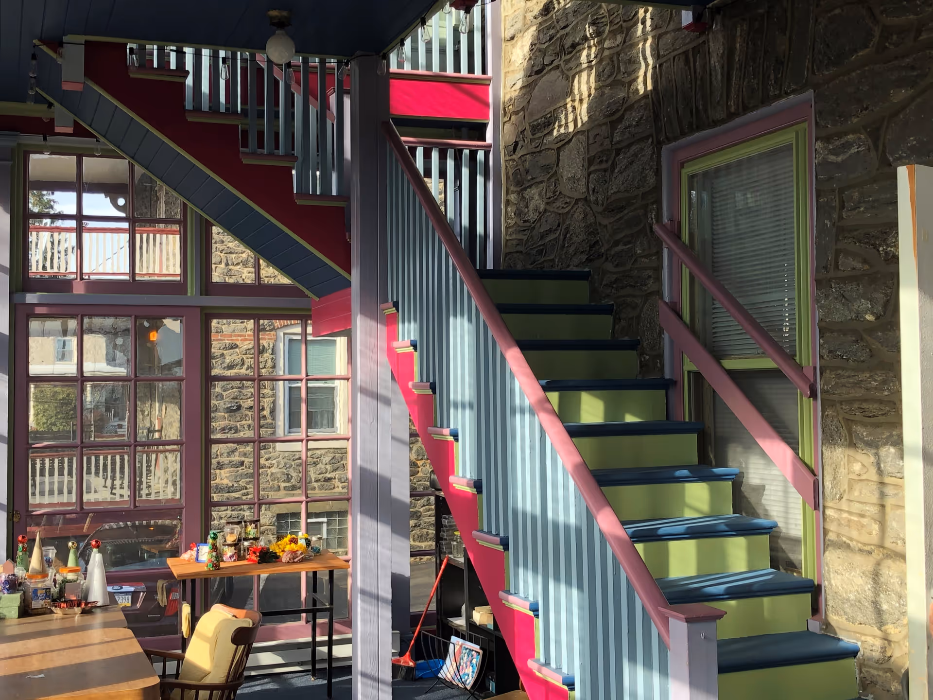 Interior view of a sunlit area with a colorful staircase featuring blue, green, and pink accents. The space has stone walls, large windows with red frames, and a wooden table with chairs and various items on it.