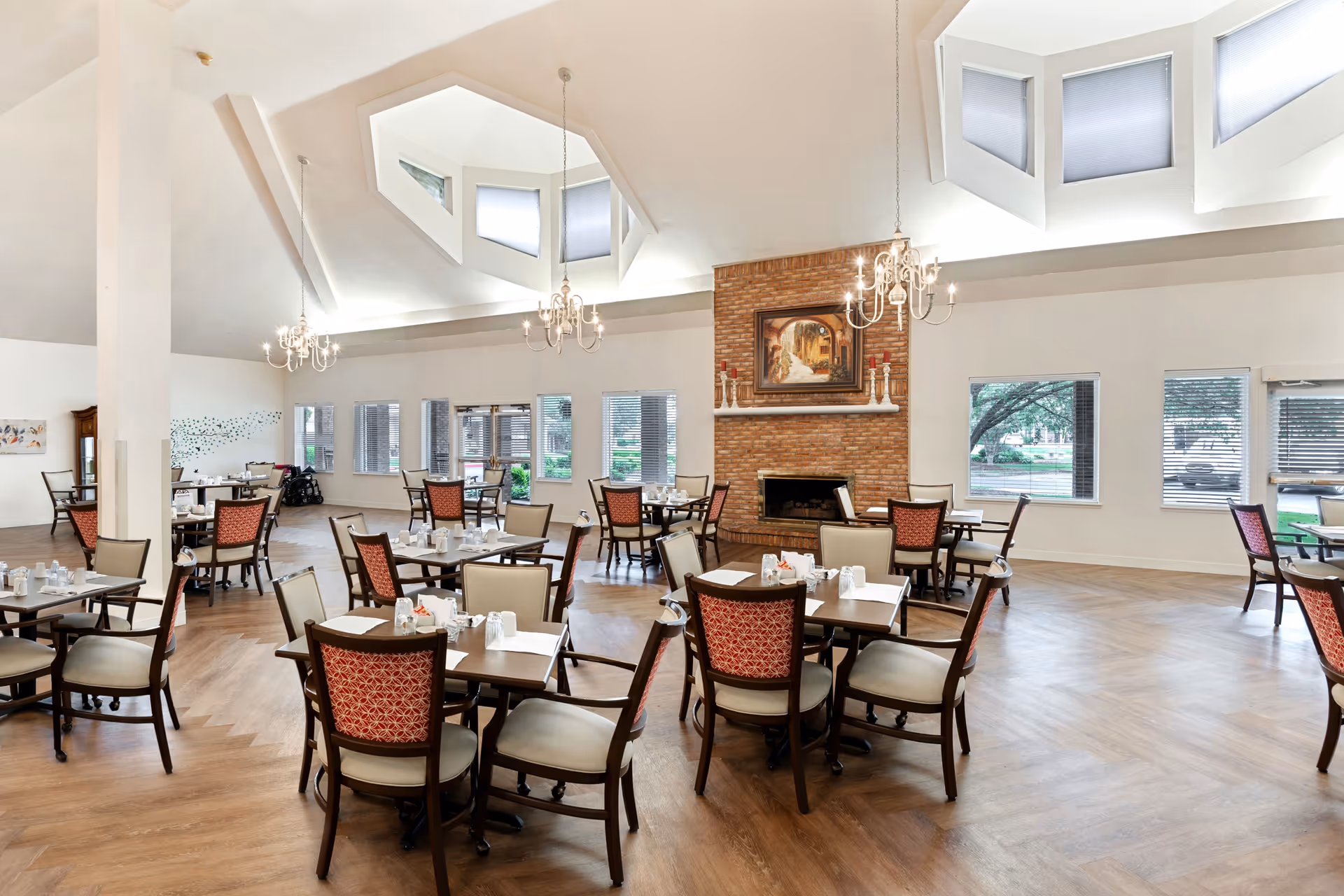 A spacious dining room with wooden flooring, featuring multiple tables set for dining. The room has large windows allowing natural light, a brick fireplace with a painting above it, and elegant chandeliers hanging from the ceiling.