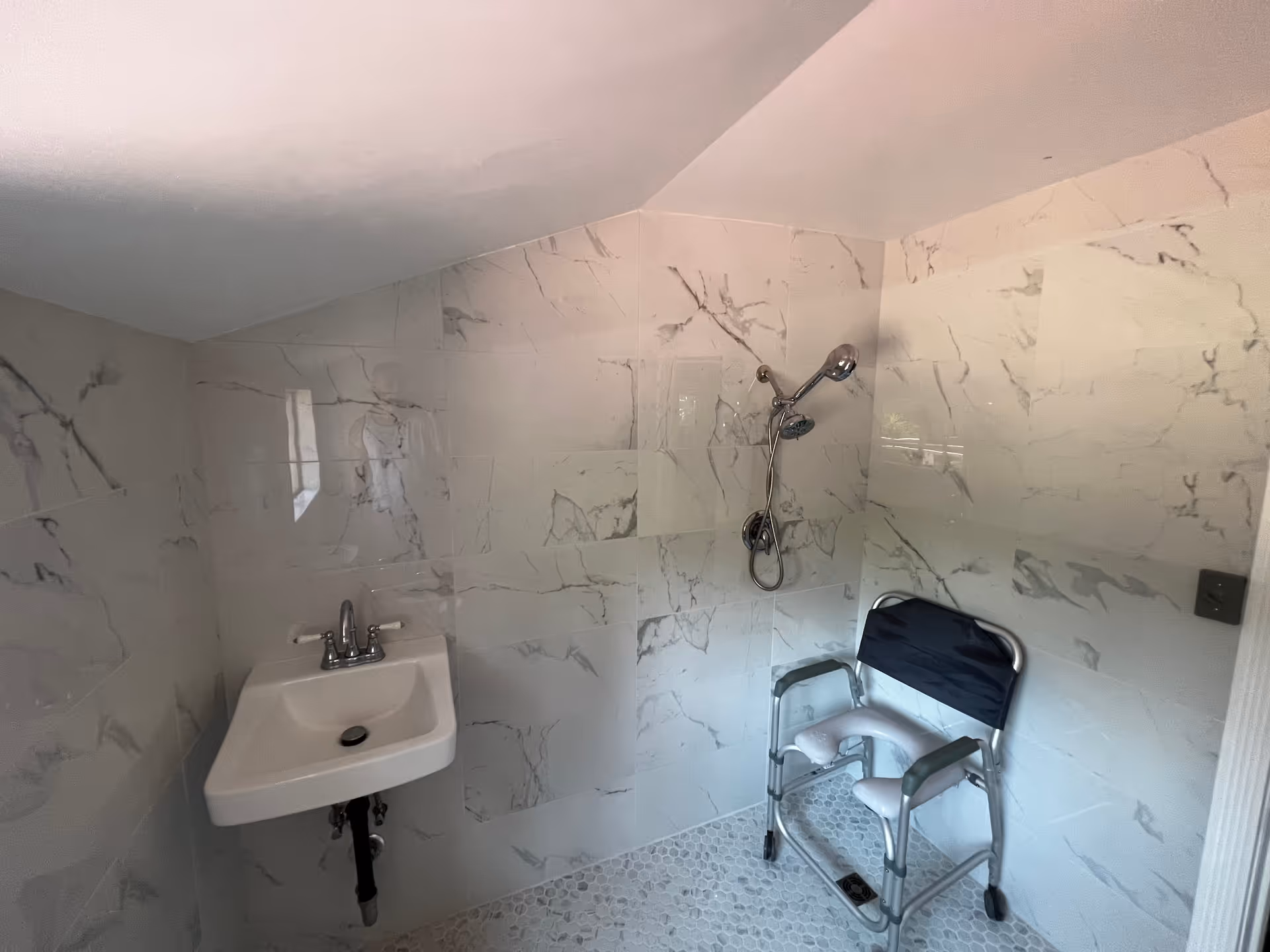 A bathroom with white marble-patterned tiled walls and a small white sink with a silver faucet. There is a shower area with a handheld showerhead mounted on the wall and a shower chair with armrests and a backrest positioned underneath it.