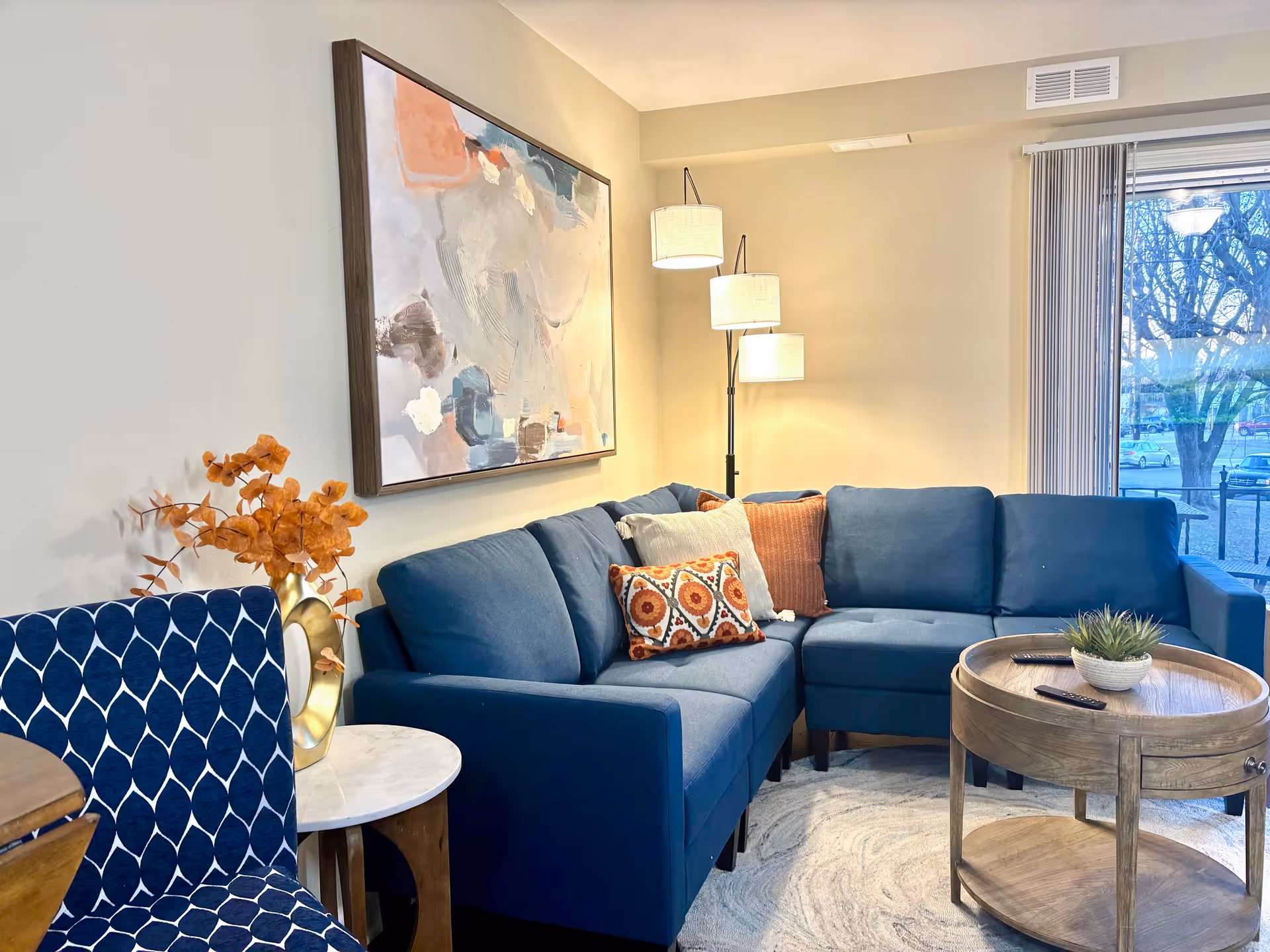 Cozy living room with a blue sectional sofa, round wooden coffee table, patterned accent chair, floor lamp, and large abstract wall art.