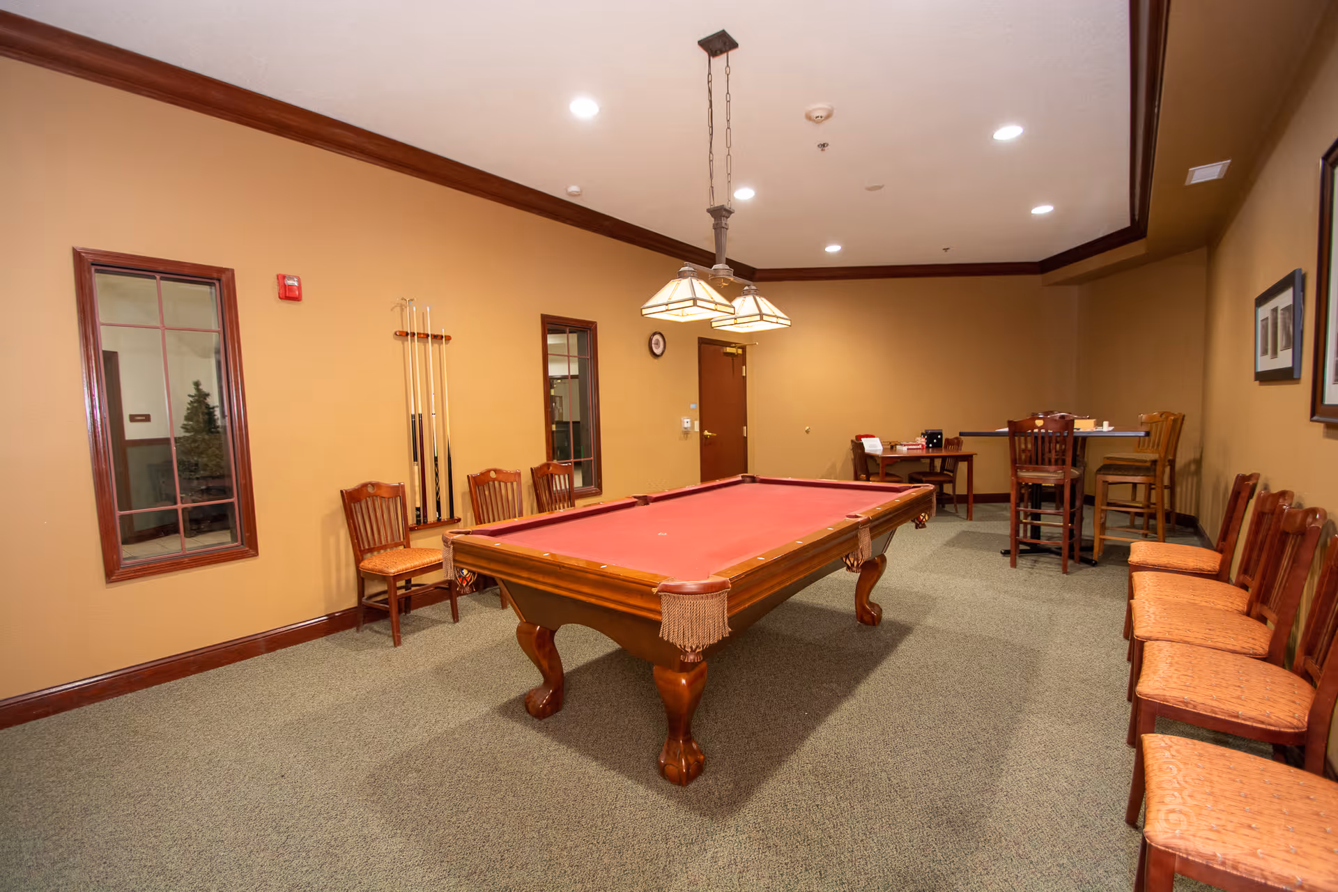 A recreation room with a red felt pool table in the center, surrounded by wooden chairs with orange cushions. The walls are painted beige with dark wood trim. There are two windows on the left wall, a clock above a door at the back, and a hanging light fixture above the pool table. In the back right corner, there is a small table with chairs.