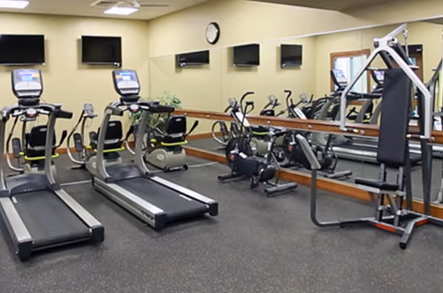 Indoor fitness room with two treadmills, two stationary bikes, a multi-function weight machine, and a large mirror on the wall reflecting the equipment. There are three wall-mounted televisions above the mirror and a clock on the wall.