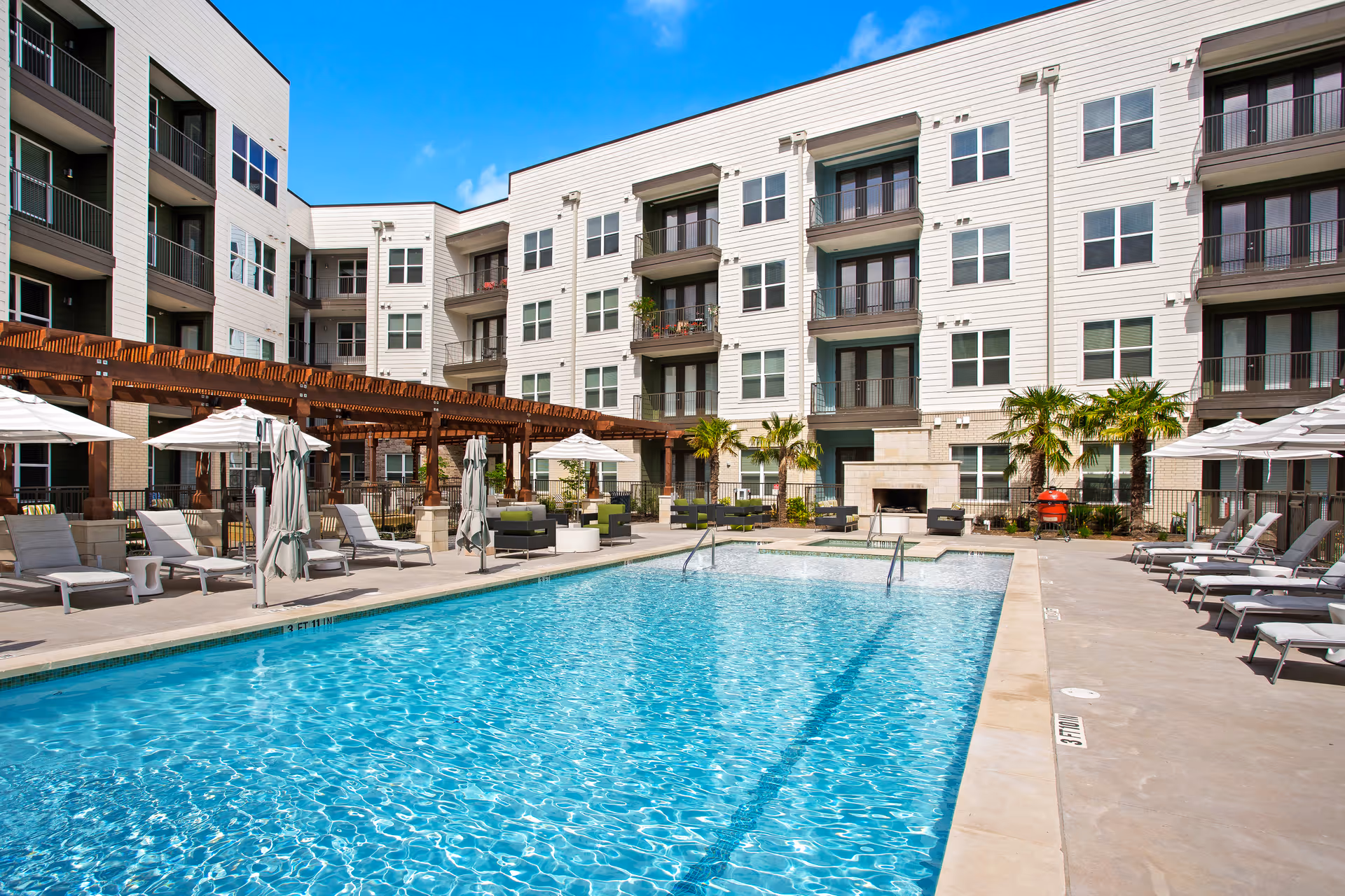 Outdoor swimming pool area at Overture Mueller facility with lounge chairs, umbrellas, palm trees, and a multi-story residential building in the background under a clear blue sky.