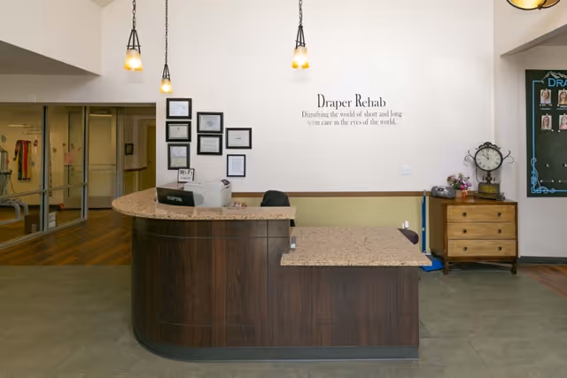 Reception desk area in Draper Rehabilitation and Care Center with a curved wooden counter, hanging pendant lights, framed certificates on the wall, and a small wooden cabinet with a clock and flowers. A sign on the wall reads 'Draper Rehab: Dignifying the world of short and long term care in the eyes of the world.'
