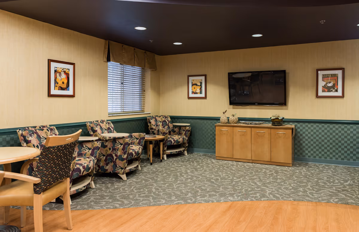 Comfortable common room with patterned armchairs and small tables facing a wall-mounted TV above a wooden cabinet.