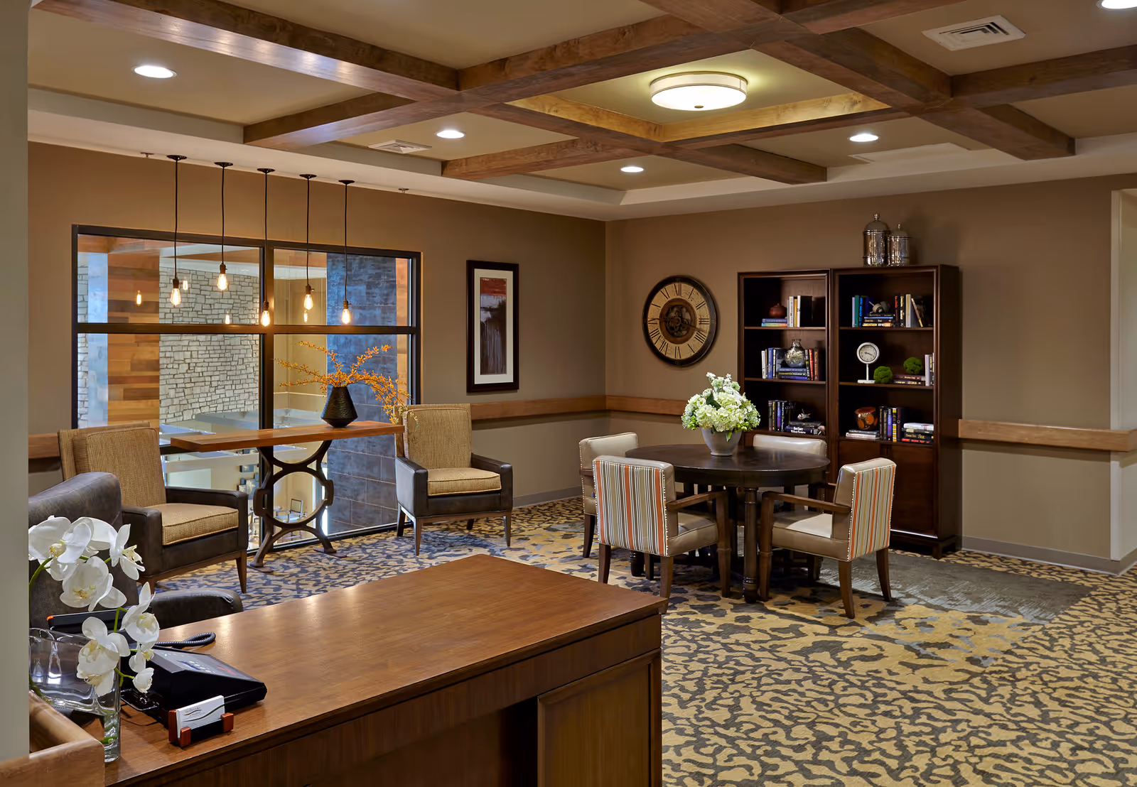 A cozy common area with a round table surrounded by four chairs, a bookshelf filled with books and decorative items, a large wall clock, and a window with hanging pendant lights. The room has a patterned carpet, wooden ceiling beams, and neutral-colored walls.