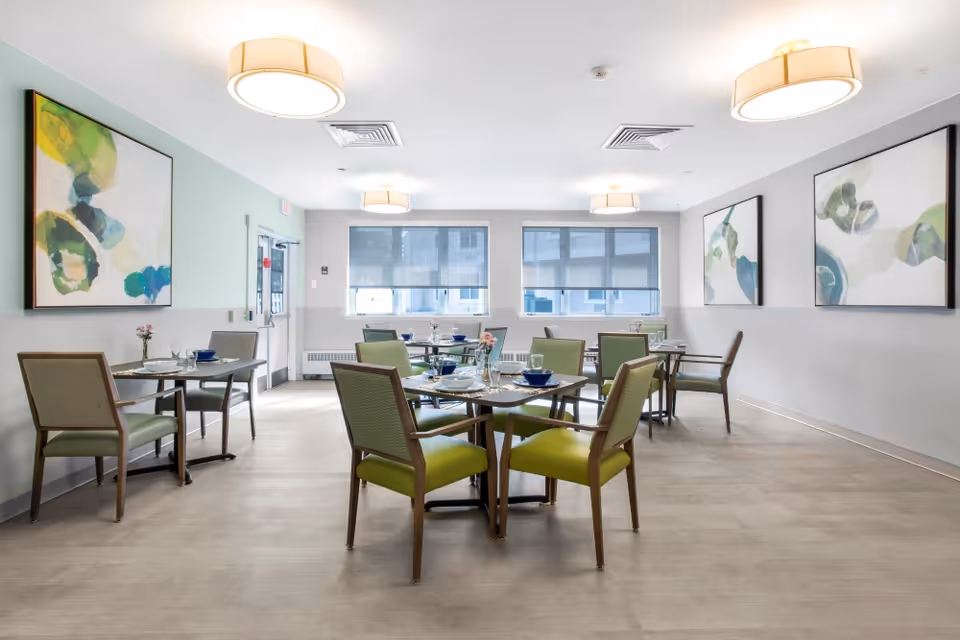 A bright dining room with several square tables set with plates, glasses, and bowls. Each table is surrounded by green cushioned chairs. The room has large windows with blinds, modern ceiling lights, and abstract artwork on the walls.