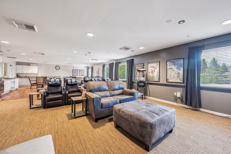 A spacious and well-lit common area in a memory care facility featuring comfortable seating including a gray sofa, matching ottoman, and black recliners. The room has large windows with dark curtains, movie posters on the gray walls, a popcorn machine, and a dining area with tables and chairs in the background. The flooring is a combination of carpet and wood.