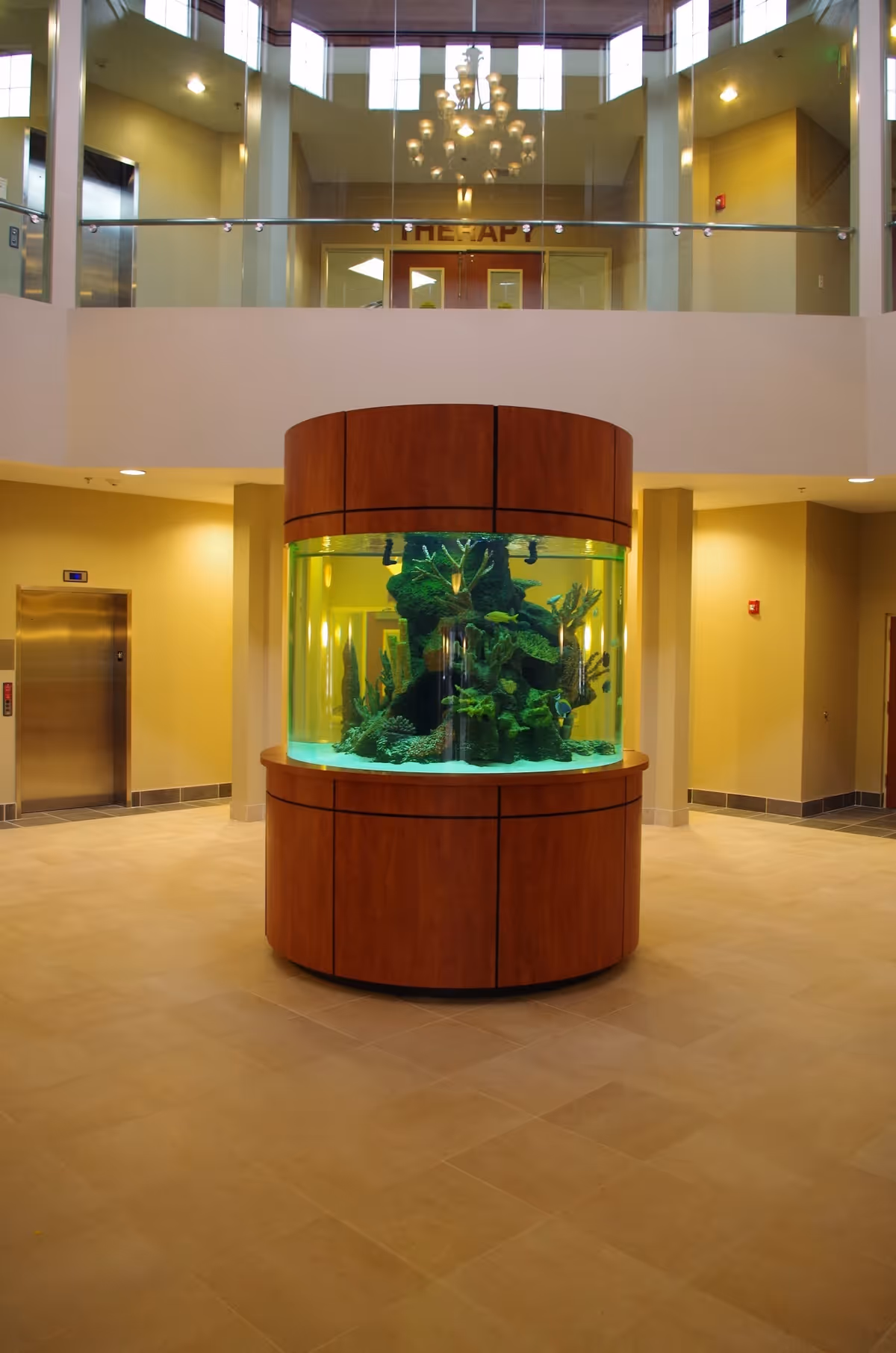A spacious lobby interior with a large round central aquarium on a wooden base beneath a mezzanine and chandelier.