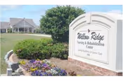 Outdoor view of Willow Ridge Nursing & Rehabilitation Center sign with a landscaped flower bed and a large tree in front of the building in the background under a blue sky.