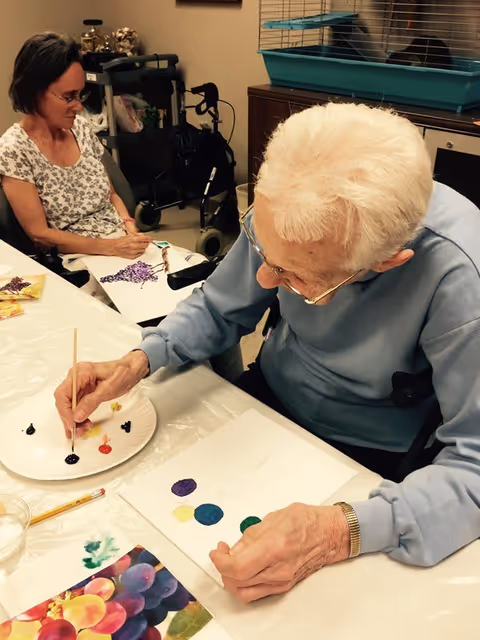 Two elderly women sitting at a table engaged in a painting activity. One woman with white hair and glasses is painting colorful circles on a piece of paper, while the other woman in a floral shirt is working on a painting of grapes. There is a small animal cage on a cabinet in the background.