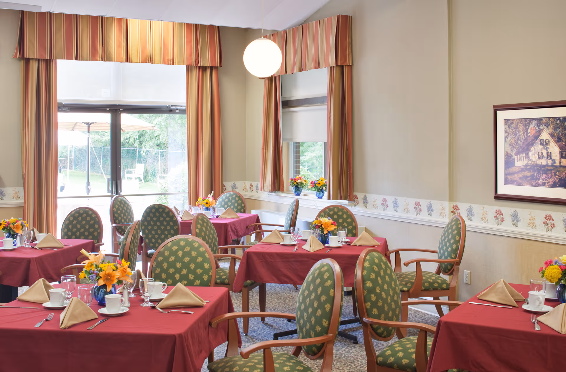A dining room with several tables covered in red tablecloths, each set with beige folded napkins, white cups, glasses, and silverware. Green cushioned chairs with wooden arms surround the tables. The room has large windows with striped curtains and a glass door leading outside. There are flower arrangements on each table and a framed picture on the wall.