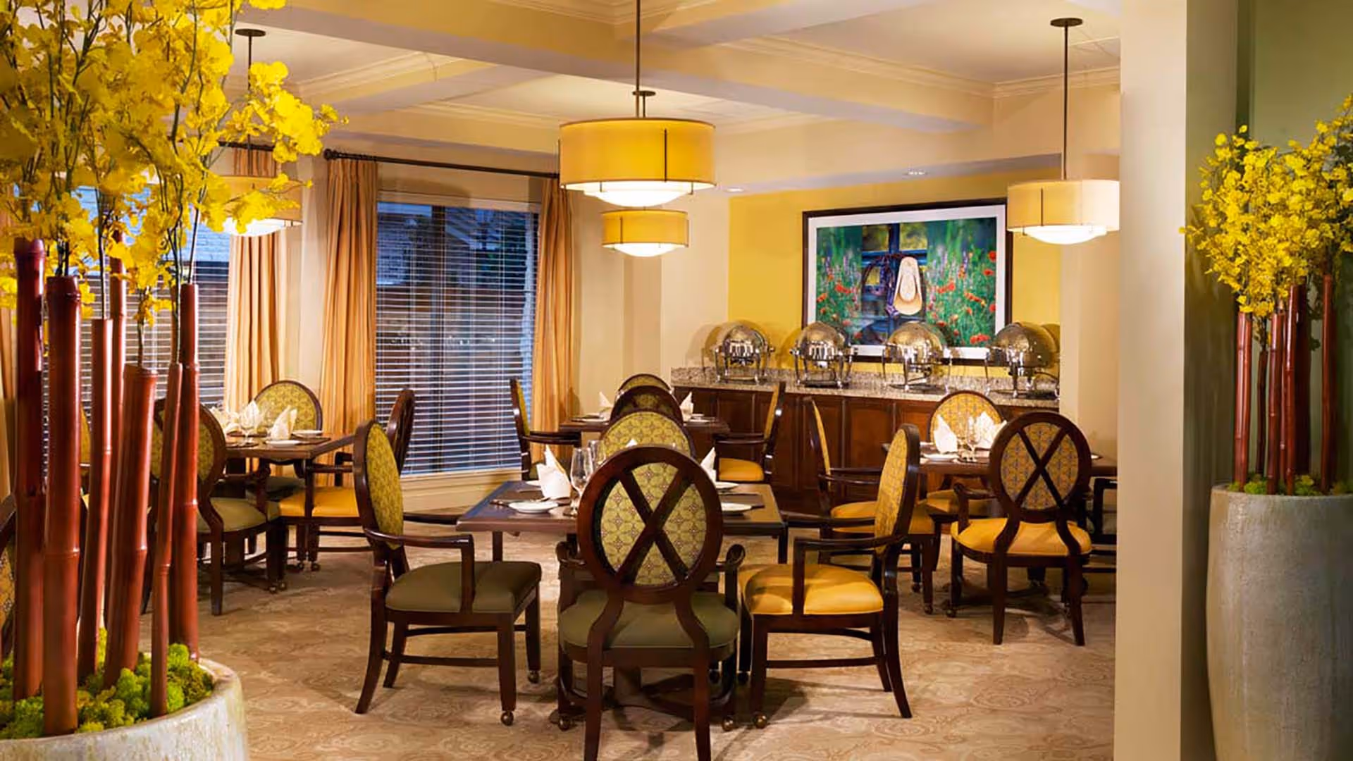 Bright dining room with round tables and upholstered chairs, buffet stations along the back wall, pendant lights, and tall yellow floral arrangements.