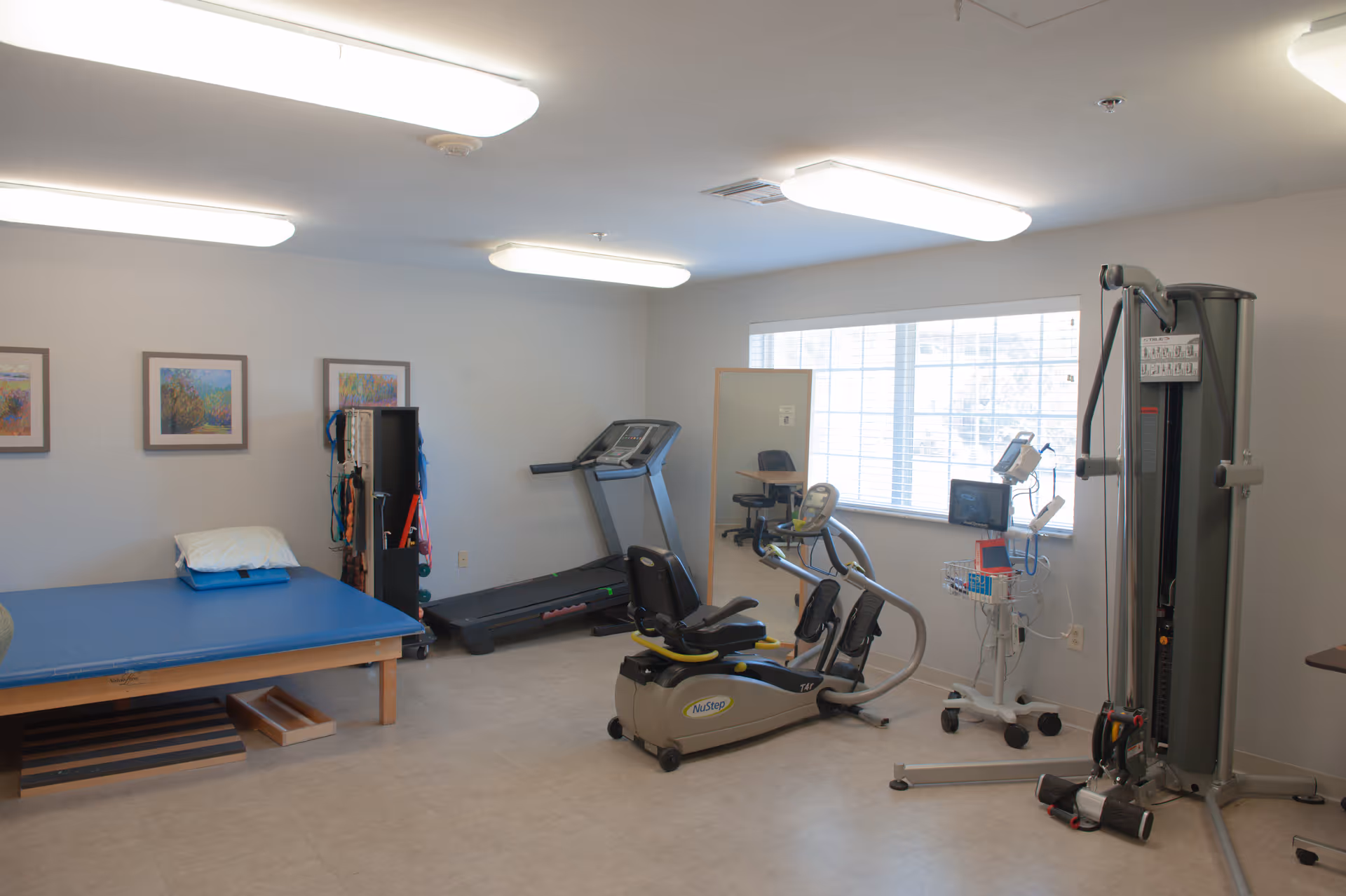 A rehabilitation room with exercise equipment including a treadmill, a recumbent bike, and a multi-function weight machine. There is also a padded therapy table with a pillow, a small shelving unit with resistance bands and other therapy tools, and a window letting in natural light. The walls are decorated with framed artwork.