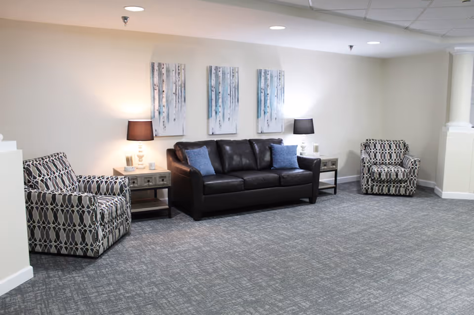 A senior living facility common area with a black leather sofa flanked by two patterned armchairs. There are two side tables with lamps on either side of the sofa, and three vertical paintings of birch trees on the wall behind the sofa. The room has light-colored walls and a gray carpeted floor.
