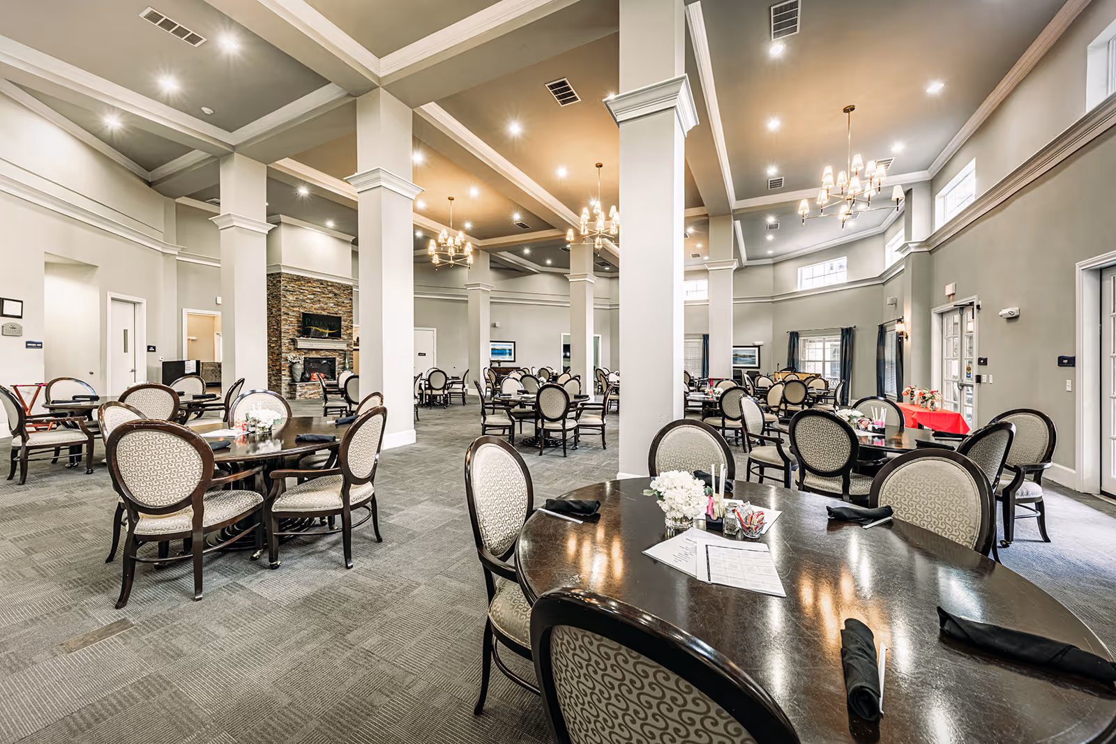 Spacious dining room with multiple round tables and cushioned chairs arranged neatly. The room features tall white columns, a high ceiling with chandeliers, large windows with curtains, and a stone fireplace in the background. The tables are set with black napkins and small floral centerpieces.