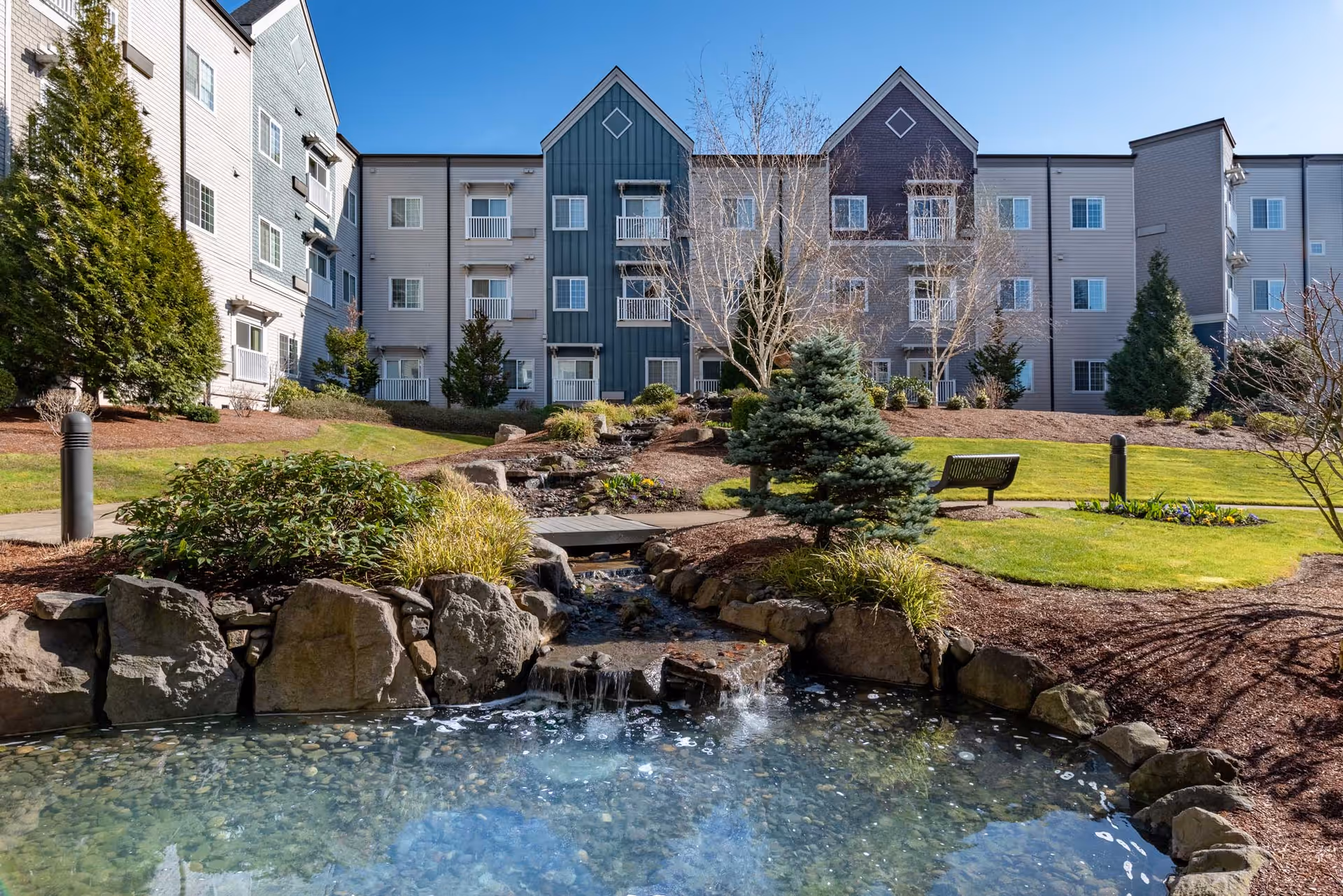 Landscaped courtyard with a small pond and waterfall in front of a multi-story senior living building.