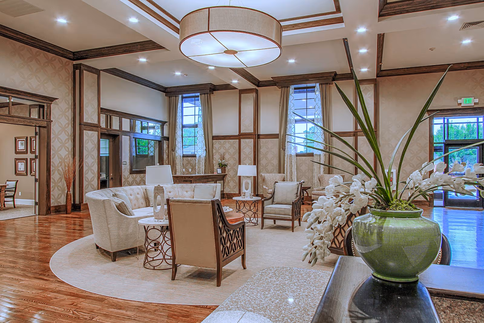 A spacious and elegant senior living facility lounge area with comfortable beige and cream sofas and armchairs arranged around small tables on a large circular rug. The room features high ceilings with recessed lighting and a large round ceiling light fixture. Large windows with curtains allow natural light to enter, and a green vase with white flowers is prominently placed on a counter in the foreground. The walls have decorative wood paneling and wallpaper, and there is a doorway leading to another room with framed pictures on the wall.