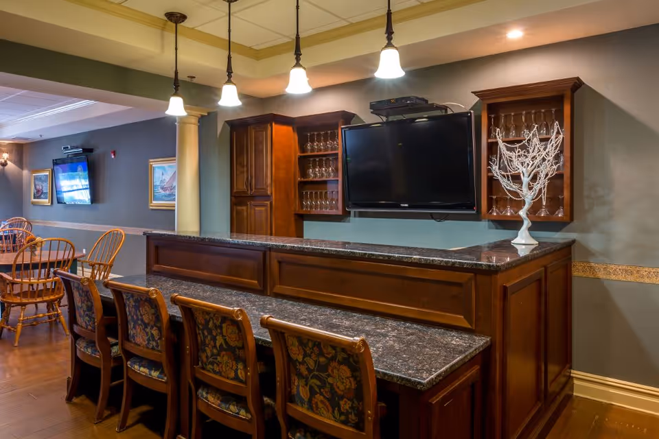 Interior view of a common area in Bellamy Fields Assisted Living featuring a wooden bar with a granite countertop, four upholstered bar stools with floral patterns, hanging pendant lights, a mounted flat-screen TV, and wooden cabinets with glassware. In the background, there are wooden dining tables and chairs, framed artwork on the walls, and another wall-mounted TV.