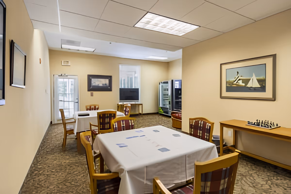 Communal dining/activity room with tables covered in white cloths, wooden chairs, framed artwork on beige walls and a vending fridge in the back.