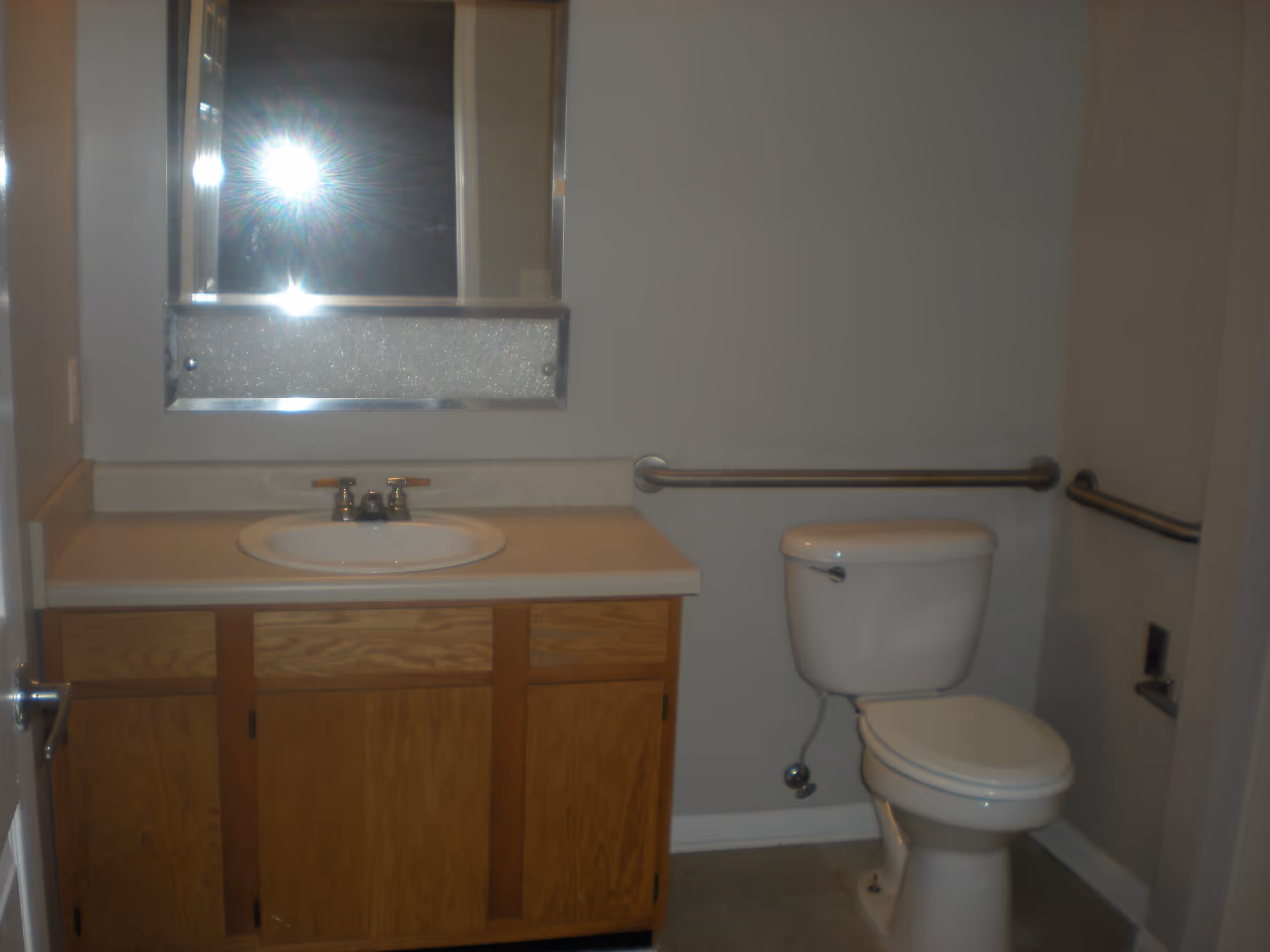 A bathroom with a wooden vanity cabinet, a countertop with a sink and faucet, a wall-mounted mirror above the sink, a toilet, and metal grab bars on the walls for support.