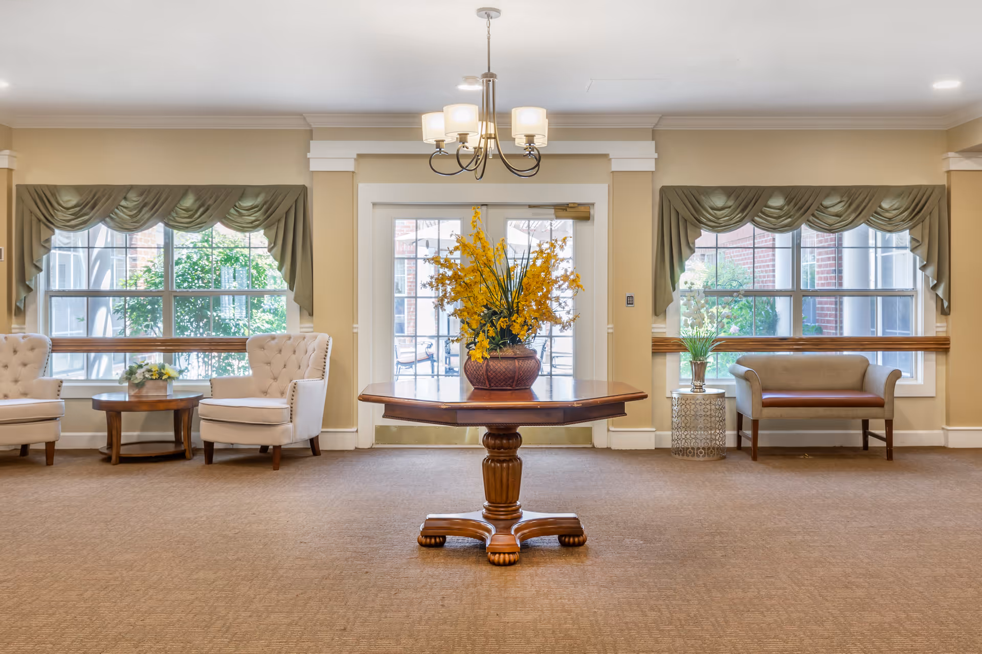 A bright and spacious common area with beige walls and carpet. In the center is a wooden table with a large floral arrangement of yellow flowers. On either side of the room are large windows with green valance curtains. To the left are two white upholstered armchairs with a small round wooden table between them holding a small flower arrangement. To the right is a small bench with a brown seat cushion and a round side table with a potted plant. A chandelier with five lights hangs from the ceiling above the central table.