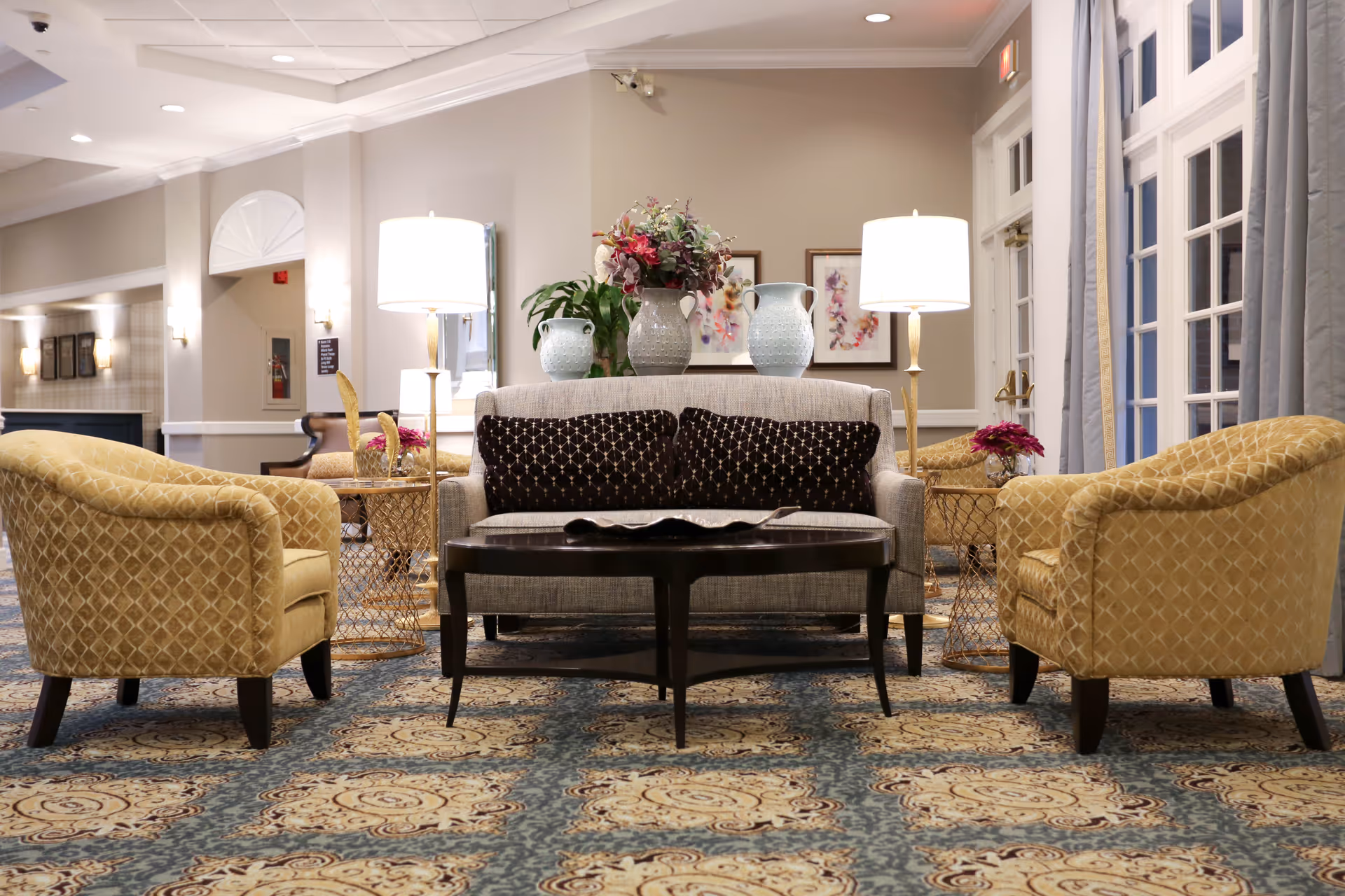 A cozy seating area in a senior living facility with a beige loveseat adorned with two dark patterned pillows, flanked by two yellow patterned armchairs. The area features a dark wooden coffee table, two side tables with lamps, and decorative vases with flowers. The room has a patterned carpet and light-colored walls with framed artwork and large windows with curtains.