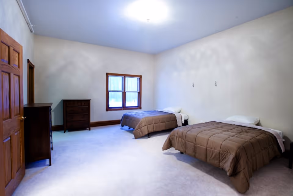A simple bedroom with two single beds covered in brown comforters, each with a white pillow. The room has beige walls, a window with wooden trim, and two wooden dressers against the wall. The floor is carpeted in light color.