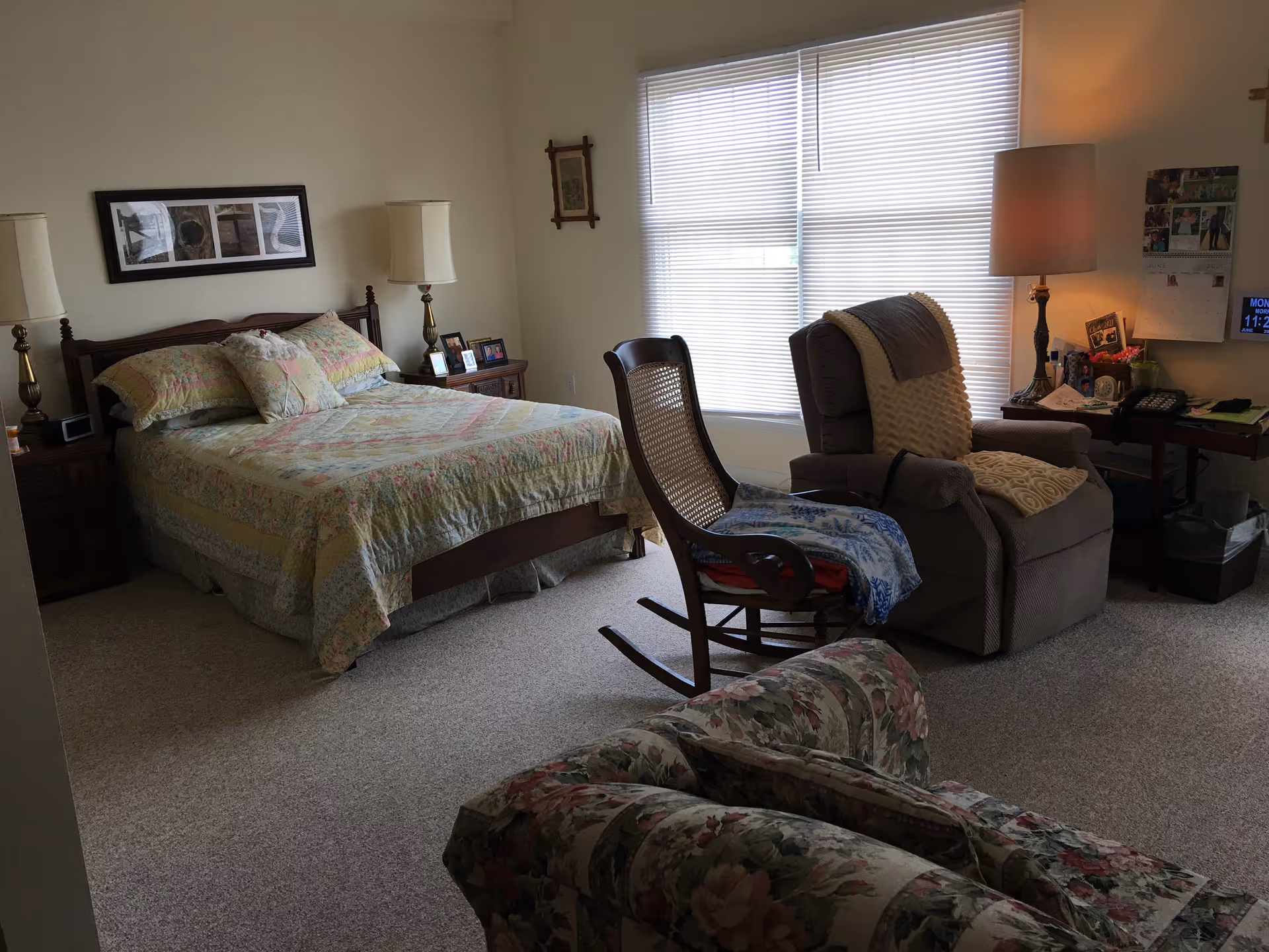 A cozy bedroom with a bed covered in a floral quilt, two bedside tables with lamps, a rocking chair, a recliner with a blanket, a floral armchair, and a window with blinds letting in natural light. There is also a small desk with a lamp, phone, and various items on it.