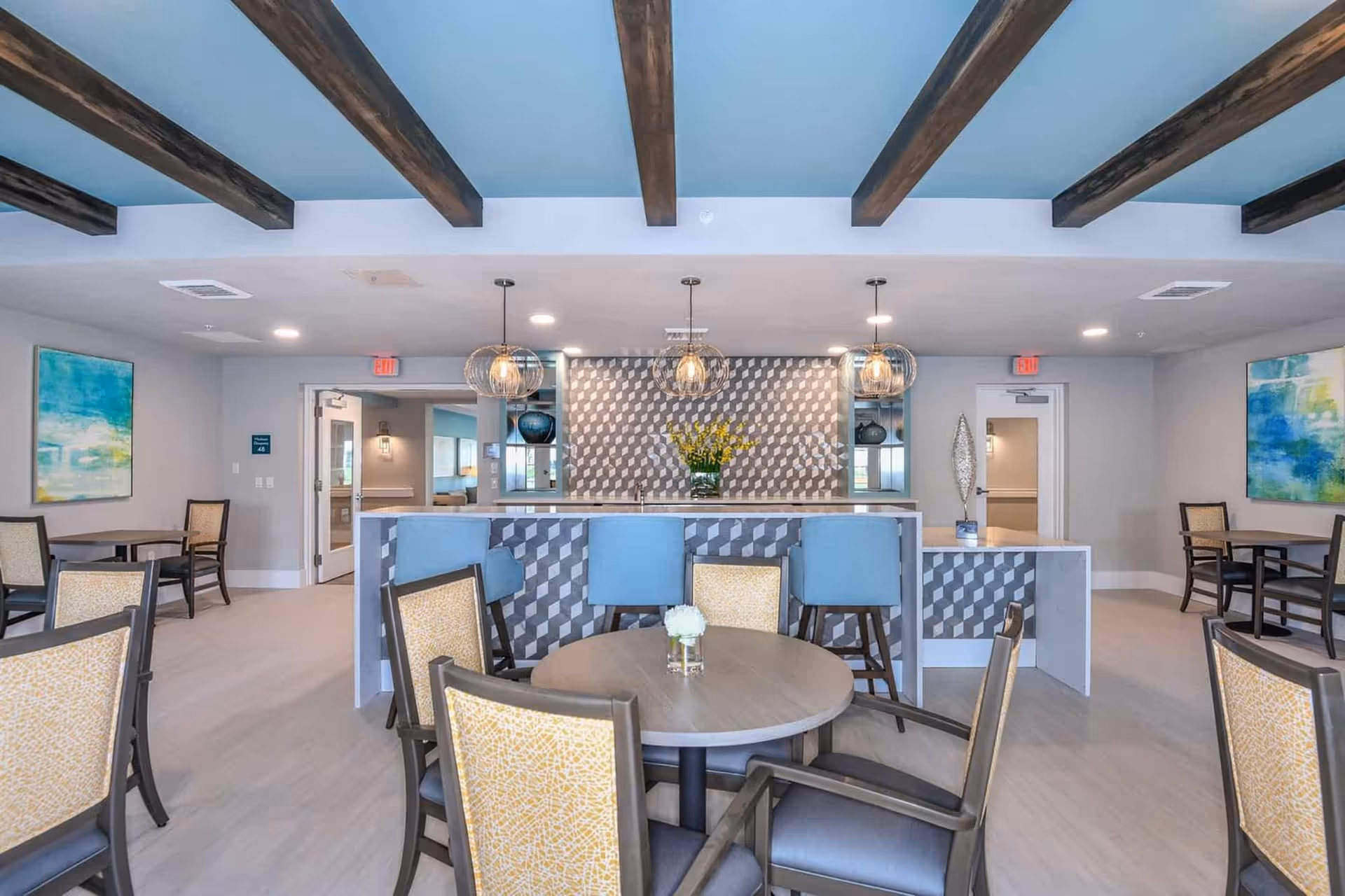 Bright communal dining area with round tables and chairs facing a decorative service counter with pendant lights and exposed wood ceiling beams.