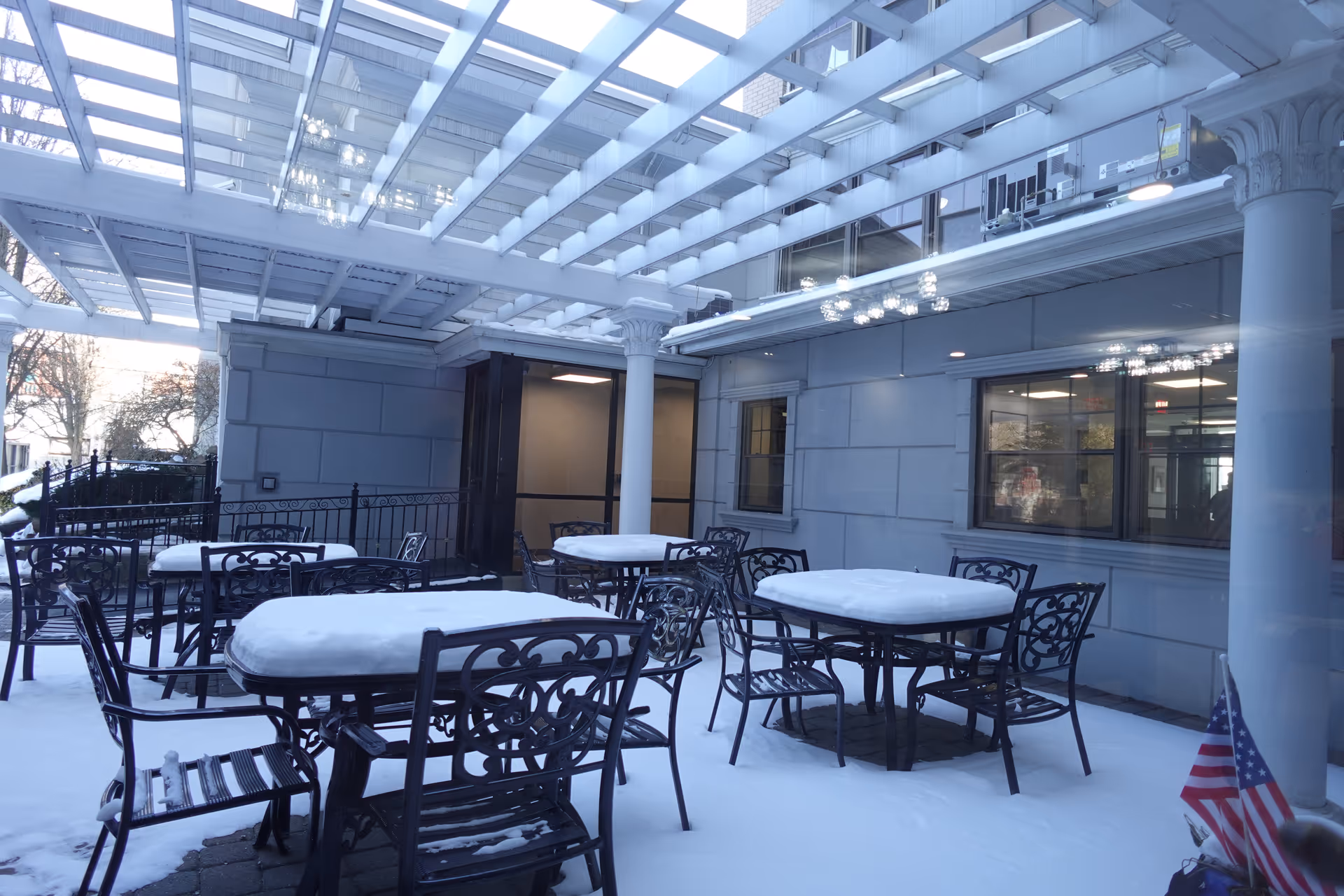 Outdoor patio area with several black metal tables and chairs covered in snow under a glass pergola attached to a building. There is an American flag in the bottom right corner and some trees visible in the background.