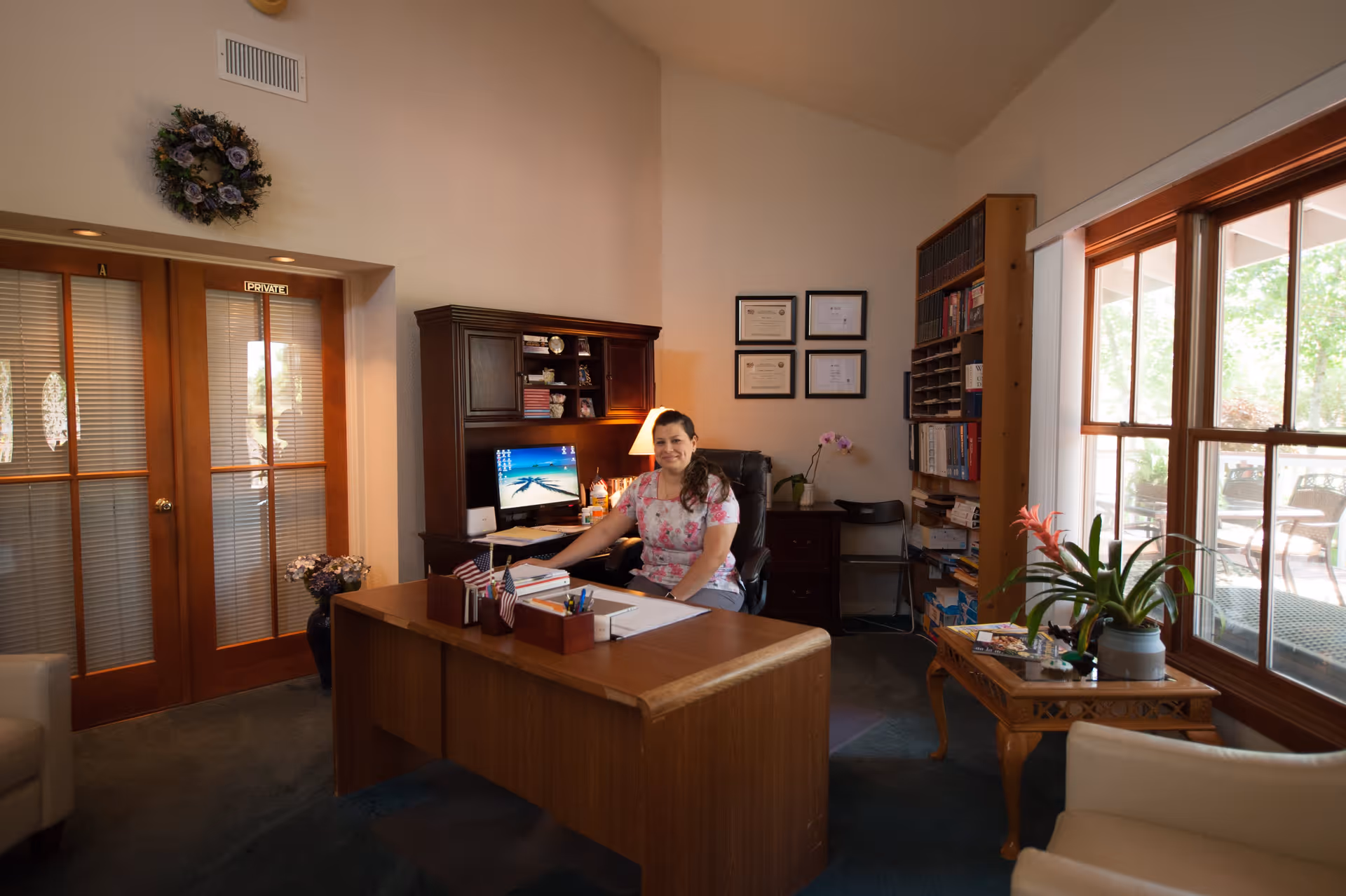 A woman sitting at a wooden desk in a warmly lit office with certificates on the wall behind her, a computer on the desk, bookshelves filled with books, and large windows showing an outdoor patio area. There are plants on a side table and a wreath hanging above double wooden doors labeled 'PRIVATE'.