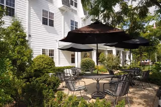 Outdoor patio with metal tables, chairs, and umbrellas beside a white multi-story senior living building surrounded by greenery.