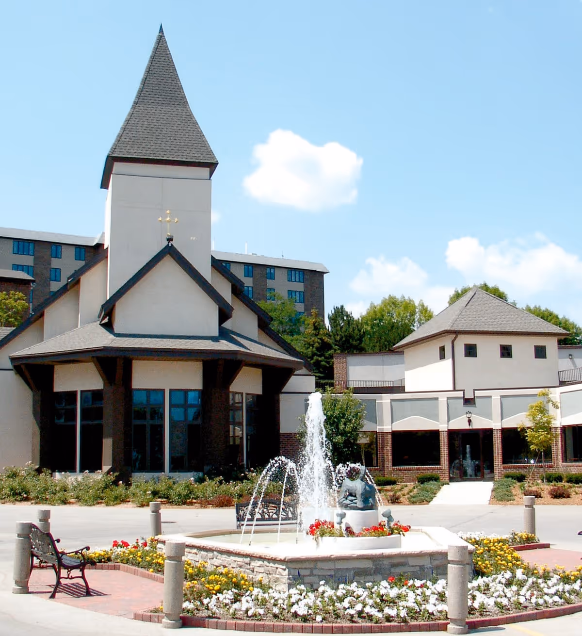 Exterior view of Ascension Living Alexian Village - Milwaukee assisted living facility featuring a building with a tall pointed roof and a cross on top, a central fountain with water jets surrounded by flowers, benches, and a clear blue sky with a few clouds.