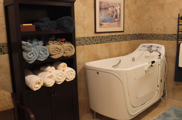 A bathroom with a walk-in bathtub featuring a door and handrails. Next to the tub is a dark wooden shelving unit stocked with neatly rolled towels in various colors. The walls are tiled with beige tiles and a decorative mosaic tile border. A framed picture hangs above the bathtub.