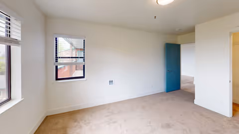 Empty room with beige carpet, white walls, two windows with blinds, and a blue door partially open leading to another room.