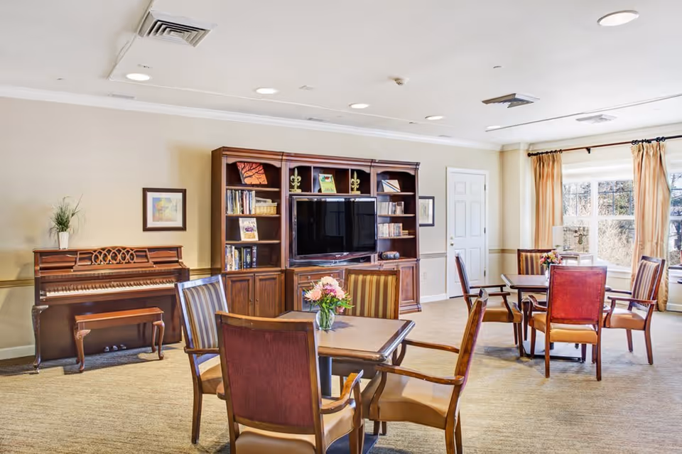 A bright and spacious common area with several wooden tables and chairs arranged for seating. There is a wooden piano with a bench on the left side of the room, and a large wooden entertainment center with a flat-screen TV and shelves filled with books and decorative items. Large windows with beige curtains allow natural light to fill the room.