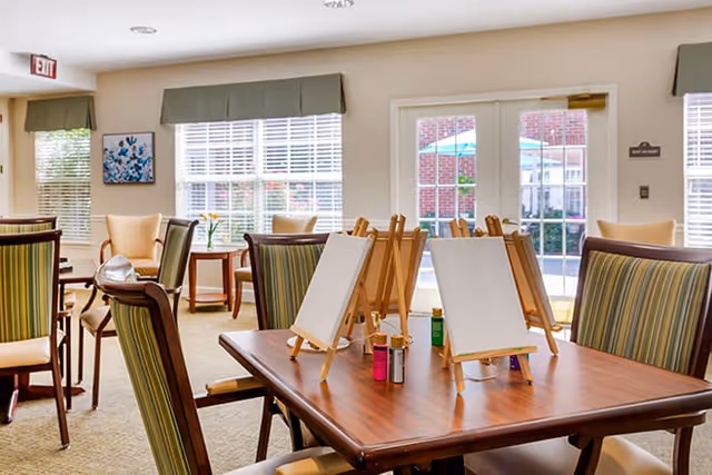 A bright communal room with tables and chairs set up with small easels and paint bottles near windows and glass doors.