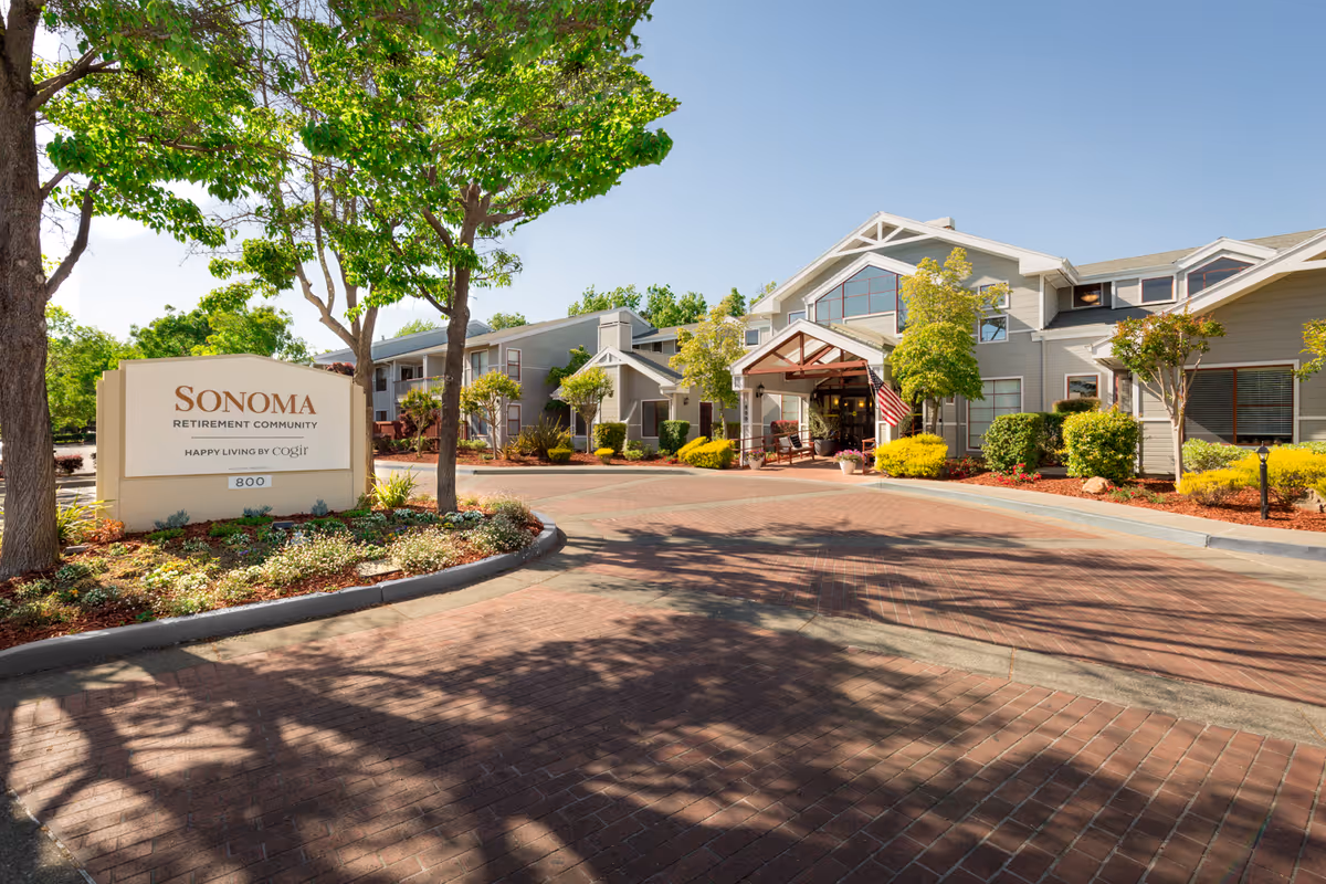 Exterior view of Sonoma Retirement Community building with a driveway, landscaped garden, trees, and a sign that reads 'Sonoma Retirement Community Happy Living by Cogir'. The building has a covered entrance with an American flag and multiple windows under a clear blue sky.