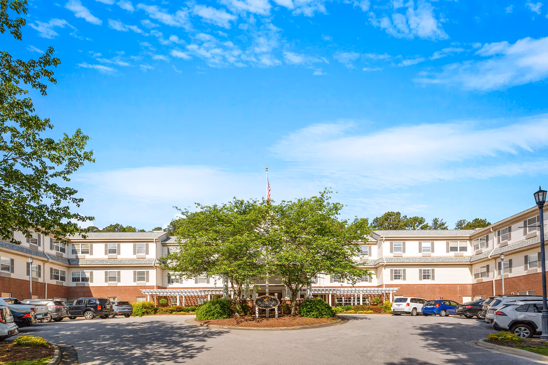 Front exterior view of Sunrise at North Hills facility with a circular driveway, parked cars, trees, and a clear blue sky.