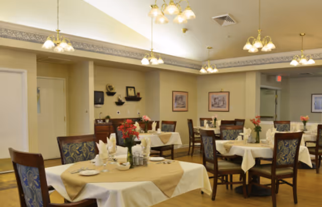 A dining room with several tables covered in white and beige tablecloths, each set with plates, glasses, napkins, and floral centerpieces. The room has wooden chairs with patterned upholstery, beige walls with framed artwork, and multiple ceiling light fixtures providing warm lighting.