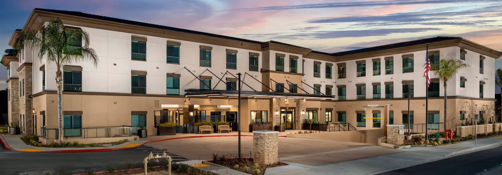 Exterior view of a three-story senior living facility building named Clearwater Newport Beach at dusk, featuring multiple windows, an entrance canopy with benches, palm trees, and an American flag.