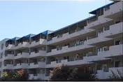 Exterior view of a multi-story residential building with multiple balconies and a few trees in the foreground under a clear sky.