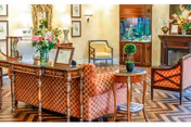 A cozy and elegantly decorated living room area with patterned upholstered chairs, a wooden table with a small plant, a large floral arrangement on a wooden console table, framed pictures on the wall, and a fish tank in the background.